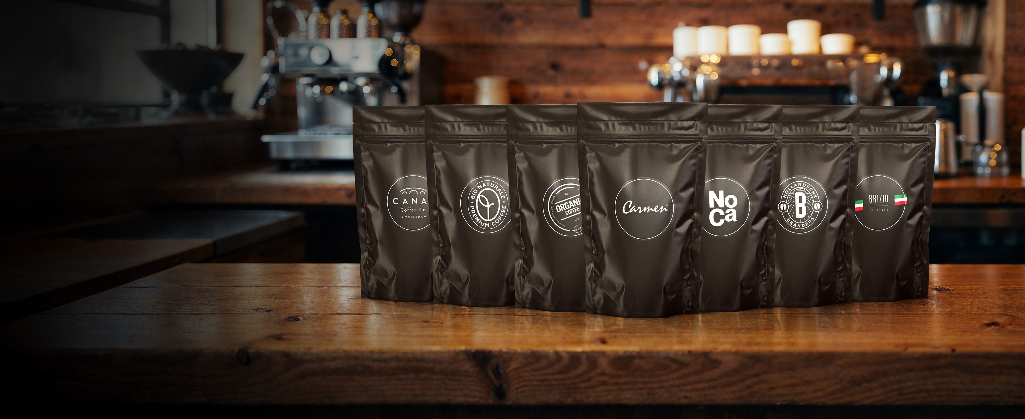 Rij zwarte testzakjes van CaféHuis met diverse koffielogo’s, gepresenteerd op een houten bar met een koffiemachine op de achtergrond.