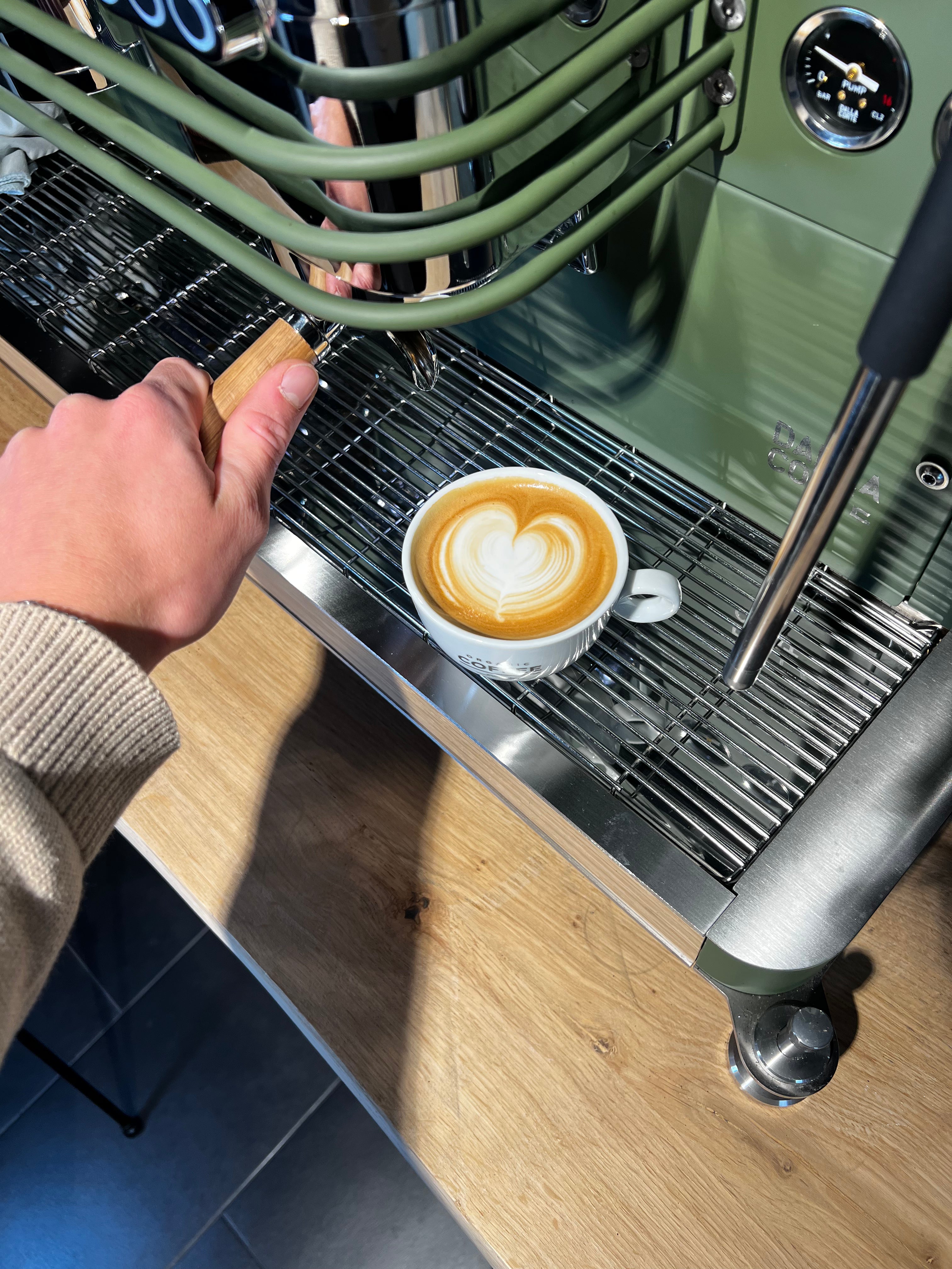 Close-up van een hand die een portafilter plaatst in een groene espressomachine; voor de machine staat een kopje cappuccino met een hartvormige latte art op het metalen rooster.